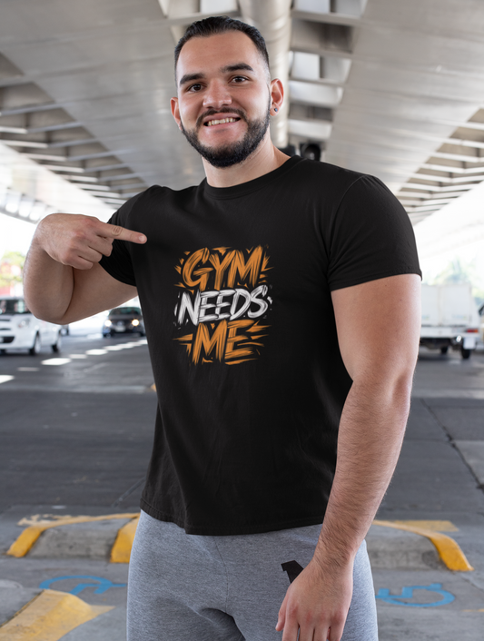 Man wearing a black t-shirt with 'GYM NEEDS ME' text, pointing at it, in an outdoor setting.